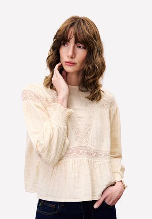 Femme aux cheveux bruns ondulés portant un blouse en dentelle crème et un jean foncé, touchant son visage et regardant légèrement sur le côté.