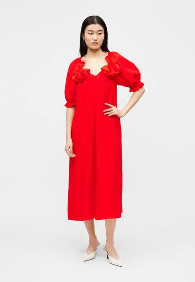 Γυναίκα στέκεται με το χέρι στη μέση, φορώντας ένα κόκκινο midi φόρεμα με φουσκωτά μανίκια και λουλουδένια διακόσμηση κοντά στη λαιμόκοψη, συνδυασμένο με λευκές γόβες.
