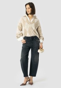 Pull en argyle beige et crème, jeans droits noirs, escarpins à imprimé léopard, tenant un sac blanc avec une poignée en anneau doré.
