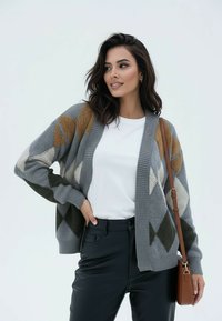 Kobieta ubrana w szary kardigan w romby, białą koszulę, czarne spodnie i brązową torebkę na ramię, pozująca z ręką na biodrze, patrząca w bok.