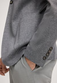 Grijze visgraat blazer met een relaxte pasvorm, vier knopen bij de mouwen en een gladde textuur, gecombineerd met lichtgrijze broek.