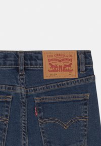 Zadní pohled na modré džíny Levi's s koženou značkovou nášivkou a šitými kapsami, přičemž na pravé kapse je červená nálepka Levi's.