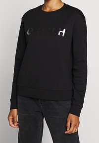 Schwarzer Sweatshirt aus weichem Material, mit Rundhalsausschnitt und langen Ärmeln. Vorne befindet sich ein reflektierendes silbernes Logo.
