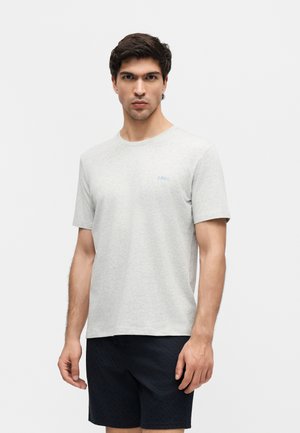 Mand iført lysegrå kortærmet t-shirt og sorte shorts står foran en ensfarvet hvid baggrund.
