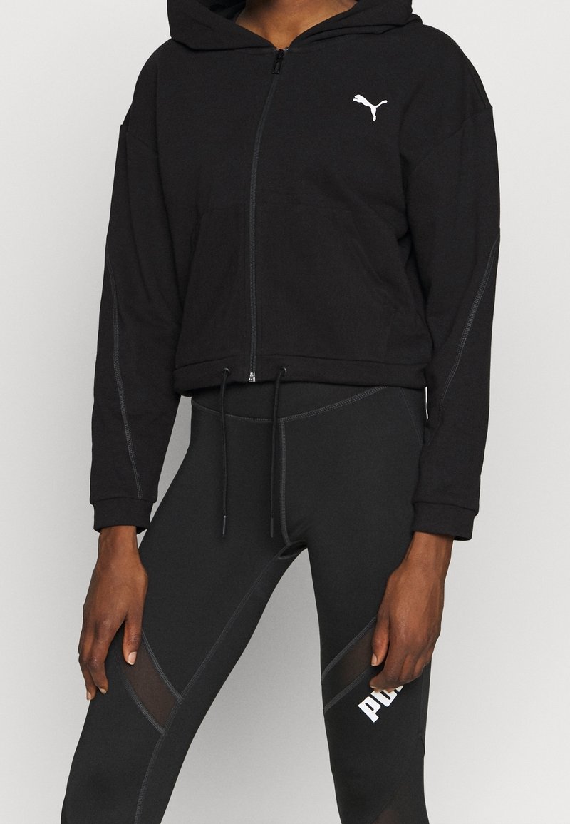 Personne portant un sweat à capuche noir zippé avec le logo Puma et des leggings noirs avec des panneaux en mesh et le texte Puma sur la cuisse.
