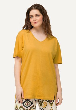 T-shirt V en col en moutarde avec des manches courtes, en tissu doux, avec des fentes latérales et une coupe décontractée, associé à un pantalon à motifs dans des couleurs contrastées.