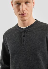 Ciemnoszara koszulka henley z długim rękawem o fakturowanym wykończeniu, z okrągłym dekoltem i trzema czarnymi guzikami przy kołnierzu.