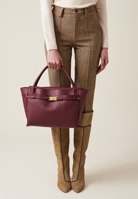 Borsa in pelle bordeaux con due manici, finitura testurizzata, accessori in metallo dorato, forma rettangolare, tenuta da una persona che indossa pantaloni a fantasia.