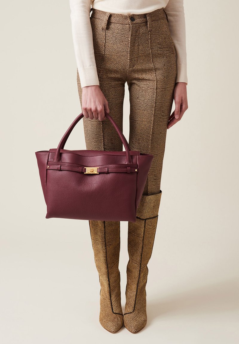 Borsa in pelle bordeaux con due manici, finitura testurizzata, accessori in metallo dorato, forma rettangolare, tenuta da una persona che indossa pantaloni a fantasia.