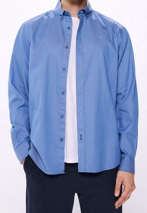 Homme portant une chemise bleu clair à manches longues déboutonnée sur un t-shirt blanc et un pantalon foncé, les mains dans les poches, debout sur fond blanc.
