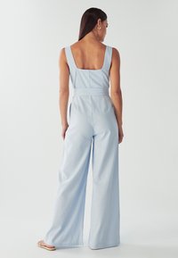 Ženska stoji obrnjenega hrbta, oblečena v svetlo modro brezrokavno širokokražno kombinezon in sandale, v preprosti ozadju.