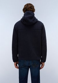 Mand med krøllet hår iført en mørk marineblå fleece-hoodie og blå jeans, står med ryggen til kameraet mod en ensfarvet baggrund.