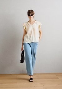 Lys gul, t-skjorte med korte ermer og volanger, kombinert med lyseblå rette jeans. Svart håndveske og svarte sandaler.