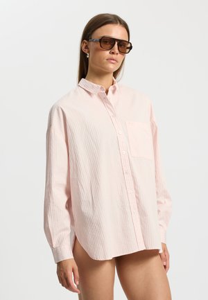 Vrouw draagt een oversized lichtroze blouse met knopen en grote bruine zonnebril tegen een witte achtergrond.