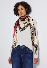 Kvinde med krøllet hår iført blå jeans, let beige langærmet sweater og et stort rødt og beige dyreprint tørklæde.