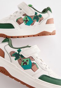 Baskets blanches avec des accents verts, comprenant des graphismes sur le thème de Minecraft. Semelle en caoutchouc texturée et bride à scratch pour un maintien sûr.
