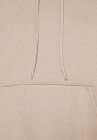 Primo piano di un felpa beige con coulisse e punte in plastica, mostrando il bordo superiore di una tasca a marsupio.