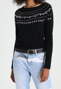Pull en tricot noir avec un col bateau, agrémenté de décorations en argent formant un motif sur la poitrine. Associé à un jean en denim bleu clair.