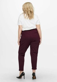 Kobieta z blond włosami, ubrana w białą bluzkę z krótkim rękawem, ciemnofioletowe spodnie i czarne szpilki, stoi na tle białym.