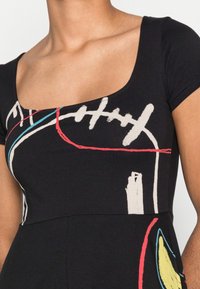 Musta lyhythihainen paita, jossa on leveä pyöreä kaula-aukko, abstrakteja värikkäitä kuvioita punaisena, valkoisena ja sinisenä, sekä tiukka siluetti.