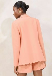 Peach-colored tailored blazer with scalloped hem, paired with coordinating shorts. Features smooth fabric and structured silhouette.