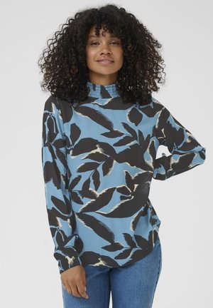 Frau mit lockigem Haar, die eine blau-schwarze, blumenmusterte Bluse mit langen Ärmeln und eine Jeans trägt, dabei mit einer Hand auf ihrer Hüfte steht.