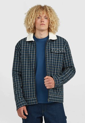 Junge Person mit lockigem blondem Haar, die eine blau karierte Jacke mit weißem Kragen und ein dunkelblaues Hemd darunter trägt.