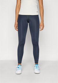 Tengerészkék sportleggings sima textúrával, testhezálló fazonnal és diszkrét oldalrészletekkel, fehér sportcipőkkel, melyeken kék akcentusok találhatók.