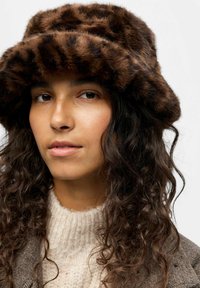 Brun faux fur bucket-hatt med en mjuk textur och horisontella ränder, bärs av en person med långt, lockigt hår.