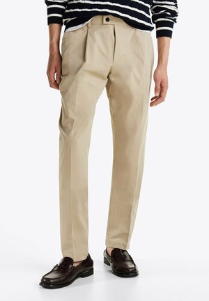 Pantaloni beige a pieghe realizzati in tessuto liscio, con gamba diritta e tasche laterali, abbinati a mocassini scuri e a un maglione a righe.
