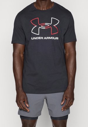 Homme portant un t-shirt Under Armour noir avec un logo blanc et rouge et un short gris, debout devant un fond clair uni.