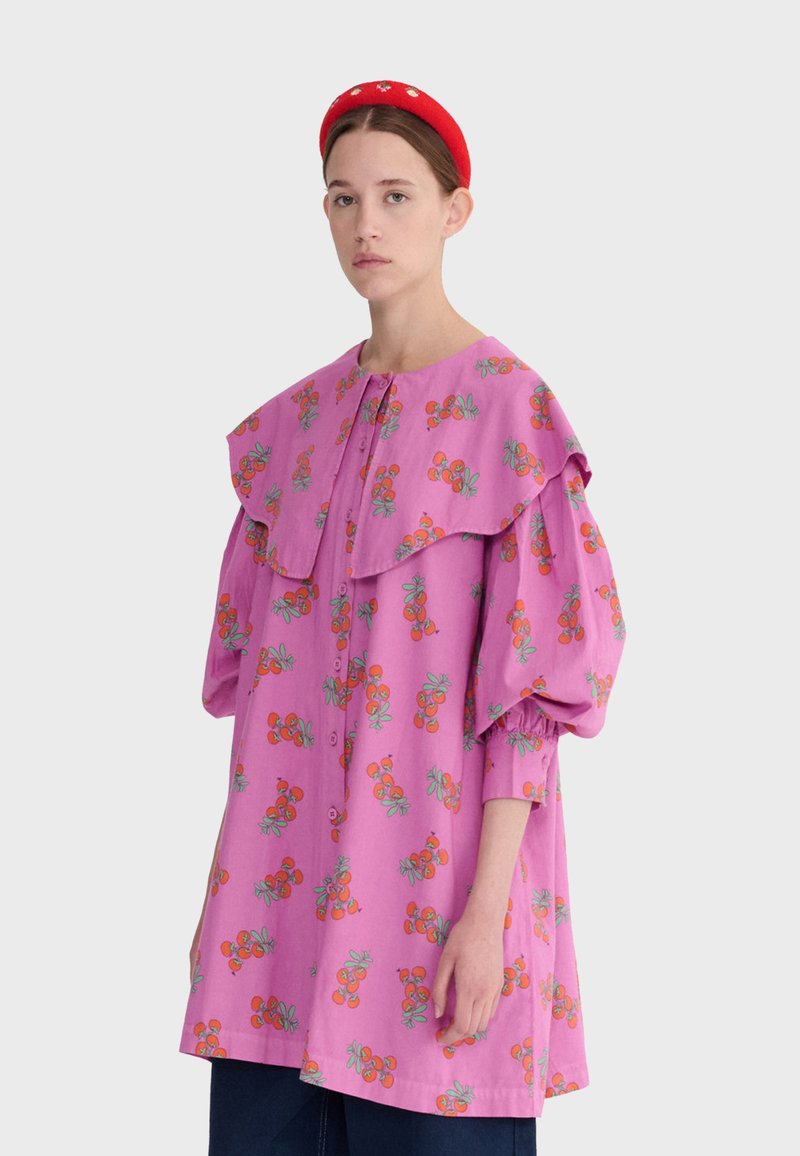 Jeune femme portant une robe rose ample avec des motifs floraux orange, un grand col, des manches bouffantes, et un serre-tête rouge.