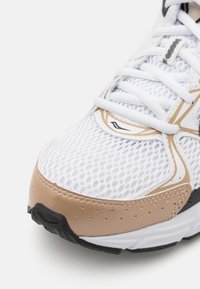 Scarpa da ginnastica bianca in rete con un accento in pelle beige, cuciture decorate e suola in gomma nera, dotata di un sistema di allacciatura prominente.