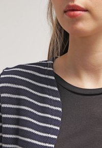 Navy and white striped knitted cardigan overlaying a black top, featuring a textured fabric and open design, highlighting horizontal lines.
