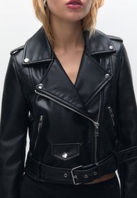 Černá motorkářská bunda z umělé kůže se stříbrnými zipy, límcem s klopami, bočními kapsami a páskem v pase. Hladká textura, přiléhavý střih.