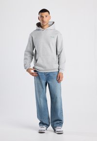Felpa grigia chiara con tasca frontale e cordini, abbinata a jeans azzurri a gamba larga e sneakers grigie, in piedi su uno sfondo bianco.