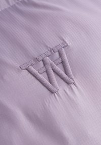Tejido púrpura con un brillo sutil y líneas verticales presenta un diseño de logo bordado en un tono de púrpura ligeramente más oscuro.