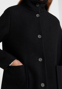 Svart ullrock med hög krage, med fem knappar och sidofickor. Tyget har en mjuk, texturerad yta.