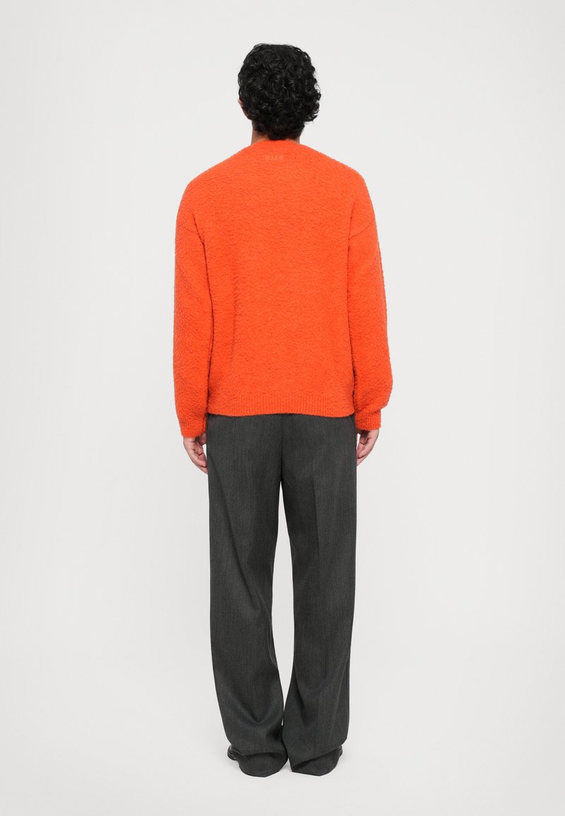Persona con capelli ricci e scuri in piedi, che indossa un maglione arancione brillante e strutturato e pantaloni grigi scuri larghi su uno sfondo semplice.