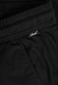 Close-up of black fabric pants with visible stitching and a small black label with white "Reell" text near the waistband.