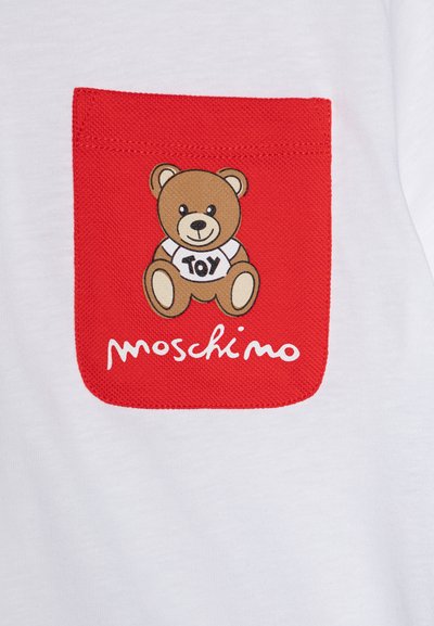 Biela tričko s červenou náprsnou kapsou s textúrovaným povrchom, zobrazuje rozprávkového medveďa s nápisom "TOY" na jeho tričku a logom "Moschino" pod ním.