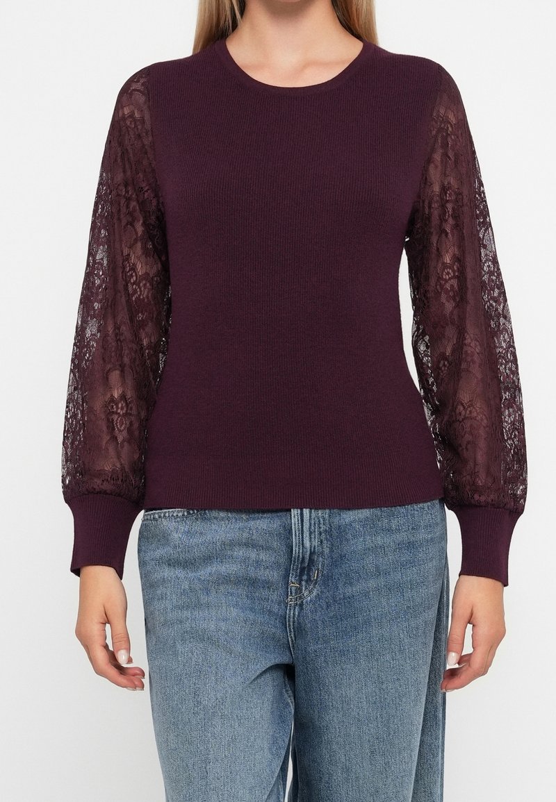 Femme portant un pull ajusté prune avec des manches évêque en dentelle transparente et des poignets boutonnés, associé à un jean en denim bleu clair.