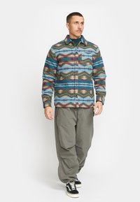 Geweven shirt in blauw, groen en bruin met een geblokt patroon, met een knoopsluiting en een borstzak, gedragen met losse olijfkleurige cargobroek en zwarte sneakers.