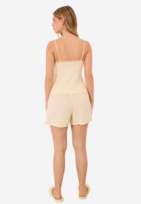 Keltainen camisole, jossa on pitsireunus, yhdistetty vaaleankeltaisiin shortseihin, joissa on pystysuuntainen raidoitus. Tossut viimeistelevät asukokonaisuuden.