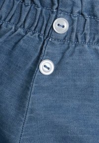 Tissu en denim bleu avec une taille froncée. Deux boutons en plastique blancs visibles, fixant le tissu avec des coutures soignées. Surface texturée.