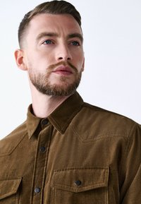 Brown corduroy shirt with a button-up front, pointed collar, twin chest pockets, and black buttons. Ribbed texture with diagonal stitching.