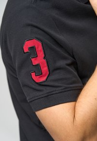 Černé polo tričko s krátkými rukávy, na levém rukávu je červeně vyšívané číslo "3". Hladká textura s přiléhavým střihem.