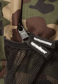 Tela de mochila de camuflaje con patrones en marrón, verde y negro. Cuenta con un bolsillo de malla negra y cremallera con detalles en blanco y etiquetas de tira.