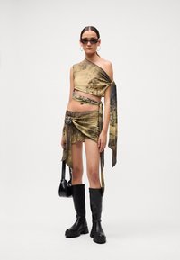 Frau in einem einseitigen Crop-Top und Minirock mit Kartenmuster, schwarzen Stiefeln bis zum Knie, Creolen, Sonnenbrille und einer schwarzen Handtasche.