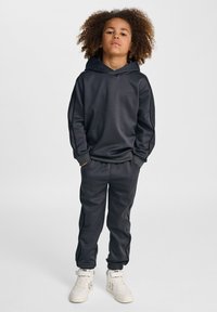 Svart träningshuva och joggingset med ribbade manschetter, sidostråk och subtil branding. Har ett mjukt, slätt tyg. Bärs med vita sneakers.